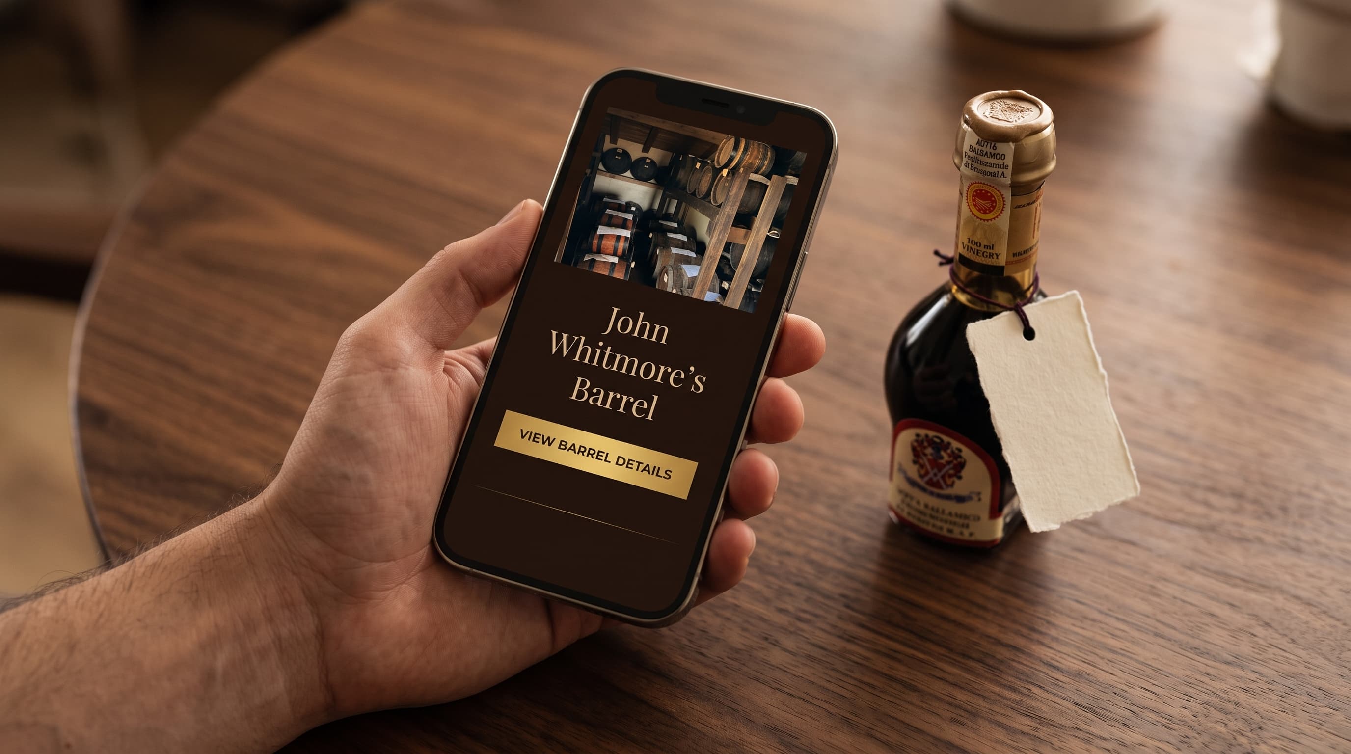 Una mano tiene un telefono aperto sulla pagina «John Whitmore's Barrel»; sul tavolo, la boccetta con il cartellino